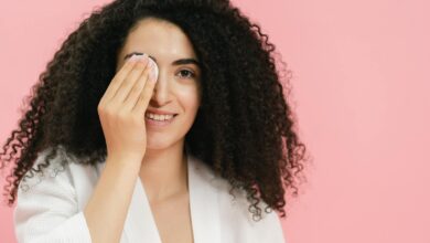 Young woman with curly hair using a cosmetic pad for skincare on a pink background.