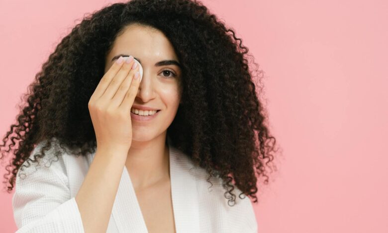 Young woman with curly hair using a cosmetic pad for skincare on a pink background.